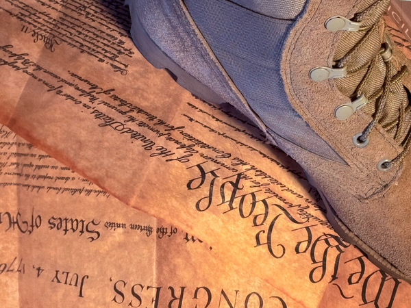 Close-up of a combat boot from above standing on the United States Constitution and the Declaration of Independence, showing upside down text from both historical documents.