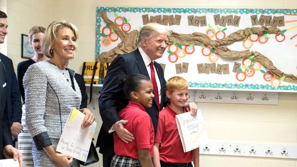Entourage, trump and two children in red school uniforms standing in a classroom, holding handmade signs, with a bulletin board behind them that reads "Orange You Glad You're in 4th Grade?"